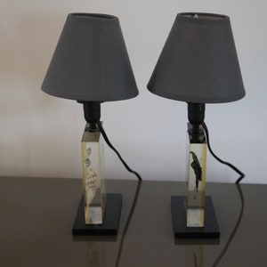 May include: Two gray lampshades with black cords and bases. Each base is made of clear acrylic with a rectangular shape and a different design inside. The bases are sitting on a dark brown surface.
