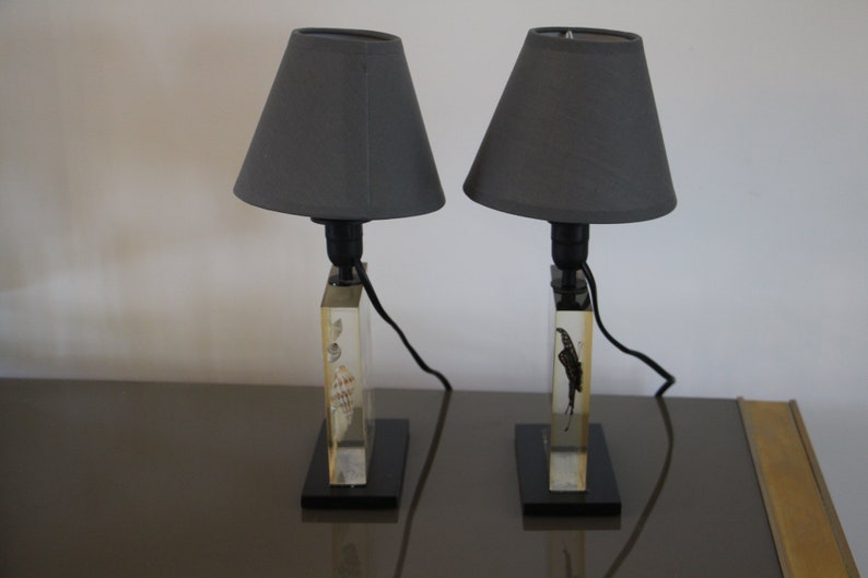 May include: Two gray lampshades with black cords and bases. Each base is made of clear acrylic with a rectangular shape and a different design inside. The bases are sitting on a dark brown surface.