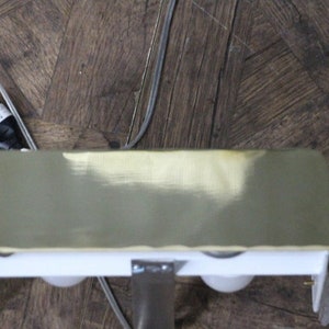 Puede incluir: Una l&aacute;mpara de color lat&oacute;n con dos bombillas blancas. La l&aacute;mpara est&aacute; montada en una superficie blanca y tiene un cable el&eacute;ctrico negro.