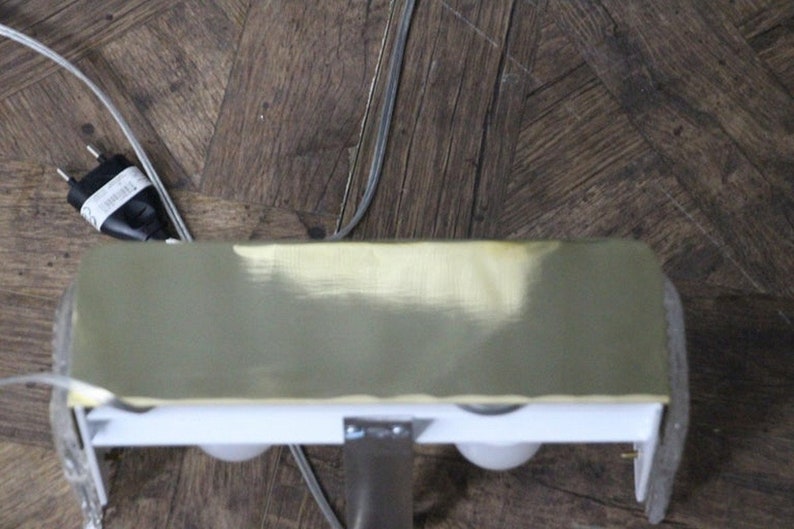 Puede incluir: Una l&aacute;mpara de color lat&oacute;n con dos bombillas blancas. La l&aacute;mpara est&aacute; montada en una superficie blanca y tiene un cable el&eacute;ctrico negro.