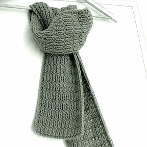 Puede incluir: Una bufanda gris tejida con un patrón texturizado cuelga de una percha blanca. La bufanda está hecha de un hilo suave y grueso y tiene forma rectangular. La bufanda es de color neutro y es adecuada para cualquier género.