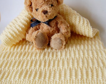 May include: A soft, cream-colored knitted baby blanket with a brown teddy bear sitting on top. The blanket has a ribbed texture and the teddy bear has a black ribbon around its neck.