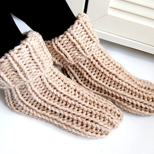 May include: A pair of beige knitted slipper socks with a ribbed texture. The socks are folded down at the top, creating a cuff.