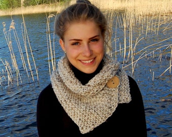 Puede incluir: Una mujer lleva una bufanda de cuello de tortuga de punto beige con un botón de madera. La bufanda se lleva sobre una camisa de cuello alto negra. La mujer está de pie frente a un cuerpo de agua con juncos en el fondo.