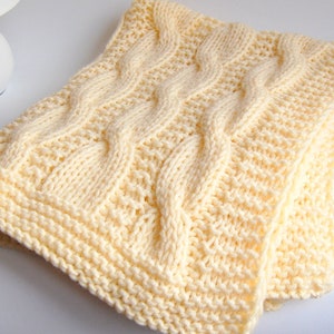 May include: A soft, cream-colored knitted blanket with a cable knit design. The blanket features a textured pattern with raised cable stitches and a ribbed border. The folded blanket is made of a thick yarn, perfect for warmth and comfort.