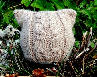 May include: A beige knitted hat with a cable knit pattern and two ear flaps. The hat is sitting on a bed of brown twigs and green grass.