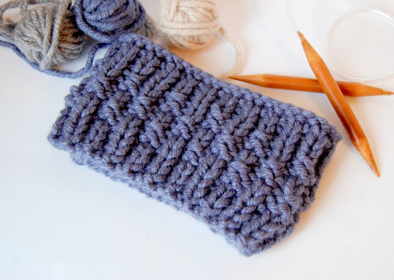 May include: A close-up of a gray knitted fabric with a textured pattern. Two wooden knitting needles are lying on a white surface.