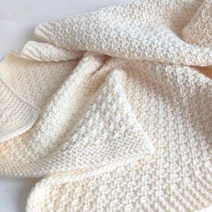 May include: A cream-colored knitted blanket featuring a textured diamond pattern throughout the fabric. The textile displays a consistent weave with visible interlocking loops, creating a soft and uniform appearance across the folded surface.