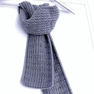 May include: A gray knitted scarf with a textured pattern, hanging on a wooden hanger. The scarf is made of a soft, thick yarn and has a rectangular shape. The scarf is displayed against a white background.