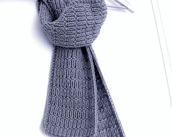 May include: A gray knitted scarf with a textured pattern, hanging on a wooden hanger. The scarf is made of a soft, thick yarn and has a rectangular shape. The scarf is displayed against a white background.