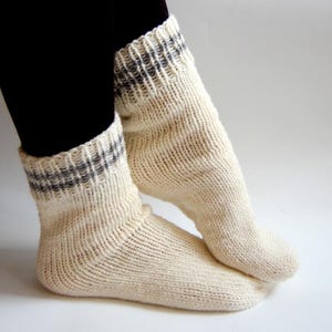 Pode incluir: Um par de meias de malha creme com punho canelado com um padrão de riscas cinza e branco. As meias são grossas e parecem quentes, adequadas para o clima frio. As meias são mostradas nos pés.