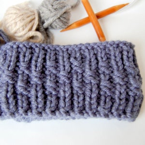May include: A close-up of a gray knitted fabric with a cable knit pattern. The fabric is lying on a white surface. There are three balls of yarn in the background, one beige, one gray, and one blue.