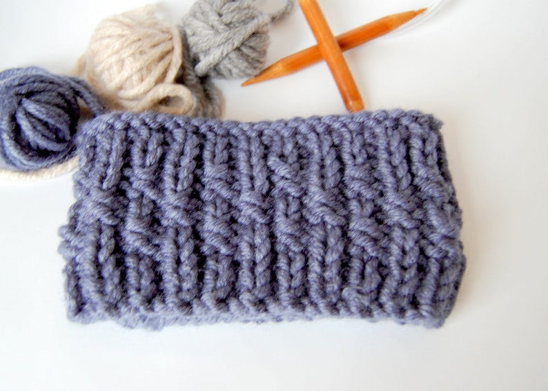 May include: A close-up of a gray knitted fabric with a cable knit pattern. The fabric is lying on a white surface. There are three balls of yarn in the background, one beige, one gray, and one blue.