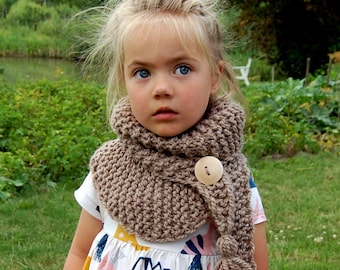 May include: A child wearing a brown knitted cowl scarf with a large wooden button closure and a tassel. The scarf is a warm, neutral color, and the child is wearing a white dress with a colorful dinosaur print.