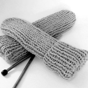 May include: A pair of gray knitted arm warmers, hand-knitted with a thick yarn, are displayed on a white surface. The arm warmers are crossed, showing the ribbed texture and the cozy, handmade quality. Knitting needles are placed beneath the arm warmers.