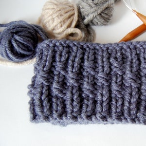 May include: A close-up of a gray knitted fabric with a cable knit pattern. The fabric is surrounded by balls of yarn in beige and gray.