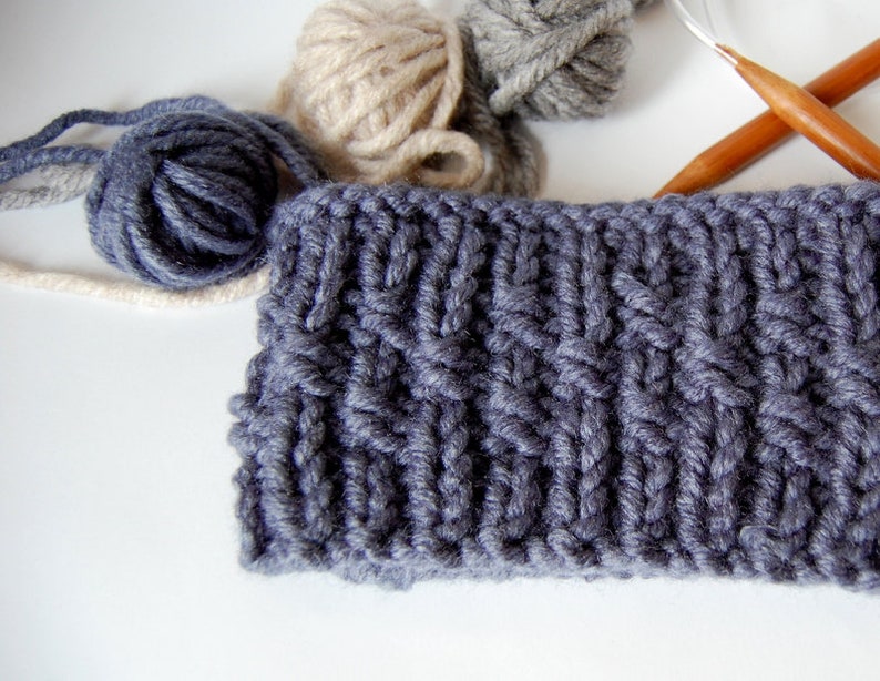 May include: A close-up of a gray knitted fabric with a cable knit pattern. The fabric is surrounded by balls of yarn in beige and gray.