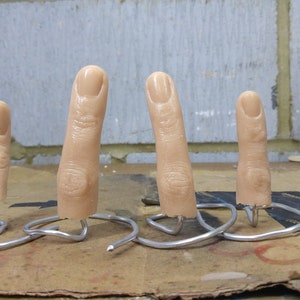 May include: Four realistic-looking silicone fingers of varying lengths are mounted on wire stands. The fingers are a pale skin tone and appear to be made of a soft, flexible material.