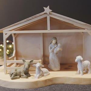 Può includere: Un presepe in legno raffigurante una stalla con una stella. All'interno ci sono statuine di Maria con Gesù bambino, un asino, un agnello e una pecora. La scena è ambientata su una base di legno.