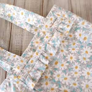 May include: A grey and white tote bag with a floral pattern of white daisies with yellow centres. The bag has a turquoise ruffle trim around the top opening and two grey straps.