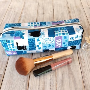Peut inclure: Trousse de maquillage rectangulaire à motifs bleus et blancs, avec fermeture éclair. Le sac présente un motif patchwork avec des motifs de chats et d'arêtes de poisson. Un pinceau de maquillage et un brillant à lèvres sont devant le sac.