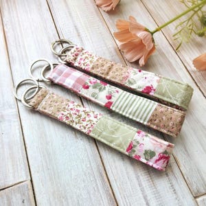 May include: Three fabric keychains with floral and patchwork patterns. The keychains feature pink, green, and beige colours. Each keychain has a silver metal ring attached for keys.
