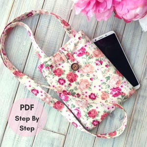 May include: A cream-colored fabric crossbody bag featuring a floral pattern with pink and red roses, a single brown wooden button closure, and a long shoulder strap. A black smartphone is tucked inside the bag, and a pink circle with the text PDF Step By Step is visible.