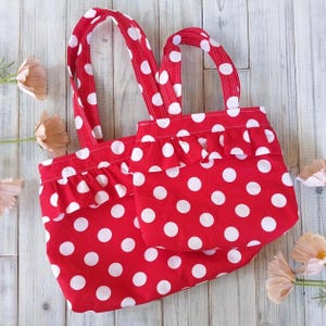 May include: A red tote bag featuring white polka dots and matching handles. The bag has a frilled trim and a smaller matching pocket. The fabric is a vibrant red with large white polka dots. The bag is designed for carrying items.