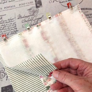 May include: Close-up of fabric pieces held together with clips. The fabric features floral and striped patterns, with a plain white layer. The background shows vintage postcard imagery. Metric measurements are not applicable.