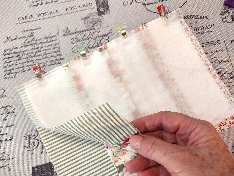 May include: Close-up of fabric pieces held together with clips. The fabric features floral and striped patterns, with a plain white layer. The background shows vintage postcard imagery. Metric measurements are not applicable.