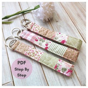 May include: Three fabric key fobs featuring patchwork designs in shades of pink, green, and beige with floral, gingham, and striped patterns. Each fob includes a silver metal split ring. A circular pink graphic contains the text PDF Step By Step.