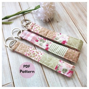 May include: Three fabric keychains with silver key rings. Each keychain is made of patchwork fabric in floral, striped, and plaid patterns, with pink, green, and beige colors. The text "PDF Pattern" is in a pink circle.