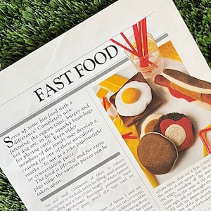 May include: A newspaper page with the headline "FAST FOOD" displays felt food toys, including a fried egg on toast, a hot dog, and a burger. The items are designed as play food or decorative items.