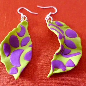 Puede incluir: Un par de pendientes con colgantes en forma de hoja verde y morada. Los pendientes están hechos de arcilla polimérica y tienen un gancho plateado.