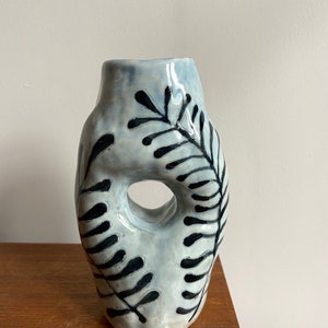 May include: A light blue ceramic vase with a unique, abstract shape and a black fern design. The vase has a hole in the side.