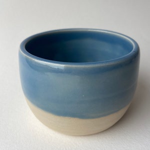 May include: A small, round ceramic bowl with a light blue glaze on the top half and a white glaze on the bottom half.