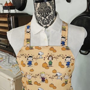 May include: A tan kitchen apron with a pattern of Peanuts characters and pumpkins. The characters are wearing fall-themed clothing and there are leaves scattered throughout the pattern.