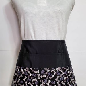 K&ouml;nnte beinhalten: Schwarze Sch&uuml;rze mit Pfoten- und Knochenmuster. Die Sch&uuml;rze hat einen schwarzen Bund und eine gro&szlig;e Tasche vorne.