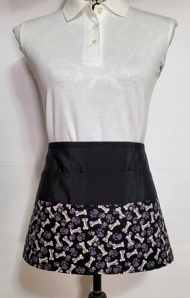 K&ouml;nnte beinhalten: Schwarze Sch&uuml;rze mit Pfoten- und Knochenmuster. Die Sch&uuml;rze hat einen schwarzen Bund und eine gro&szlig;e Tasche vorne.