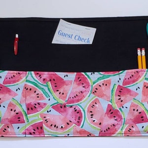 May include: A black fabric apron with a watermelon print. The apron has multiple pockets and ties at the waist. The pockets contain a pair of scissors, pencils, markers, and a guest check.