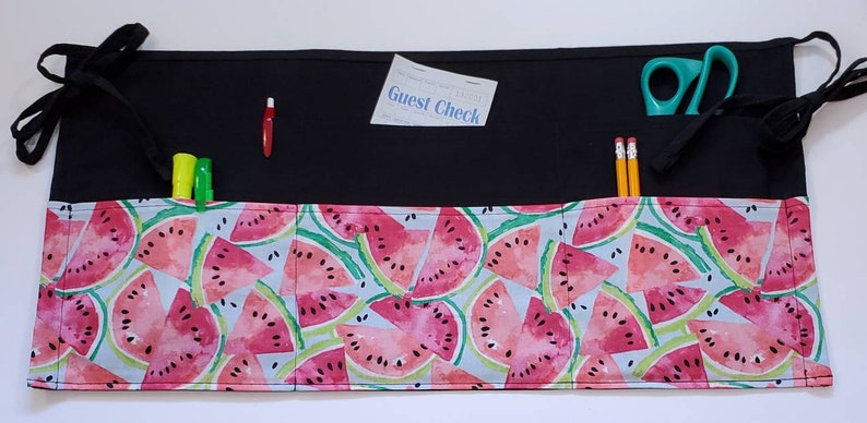 May include: A black fabric apron with a watermelon print. The apron has multiple pockets and ties at the waist. The pockets contain a pair of scissors, pencils, markers, and a guest check.