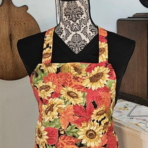 May include: A black apron with adjustable straps features a colorful sunflower and autumn leaf print. The print includes red, orange, yellow, and green leaves and sunflowers.