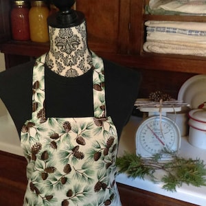 Op de afbeelding: Een witte schort met een print van dennenappels en groenblijvende planten. De schort heeft verstelbare banden en wordt getoond op een mannequin.