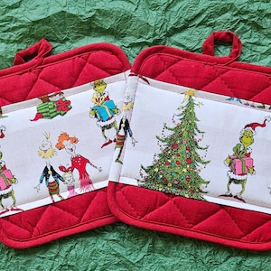 May include: Two red quilted pot holders with white fabric inserts featuring a Christmas tree and characters from the Grinch movie. The fabric is printed with a green Grinch, a Christmas tree, and other characters.