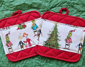 May include: Two red quilted pot holders with white fabric inserts featuring a Christmas tree and characters from the Grinch movie. The fabric is printed with a green Grinch, a Christmas tree, and other characters.