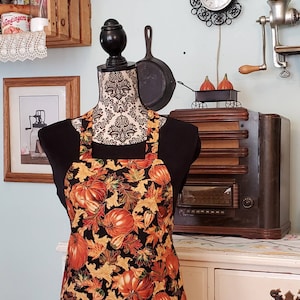 May include: A black apron with a fall-themed print of orange pumpkins and yellow leaves on a black background.