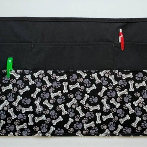 K&ouml;nnte beinhalten: Schwarze Sch&uuml;rze mit mehreren Taschen und einem Muster aus Hundeknochen und Pfoten. Die Sch&uuml;rze hat zwei verstellbare B&auml;nder.