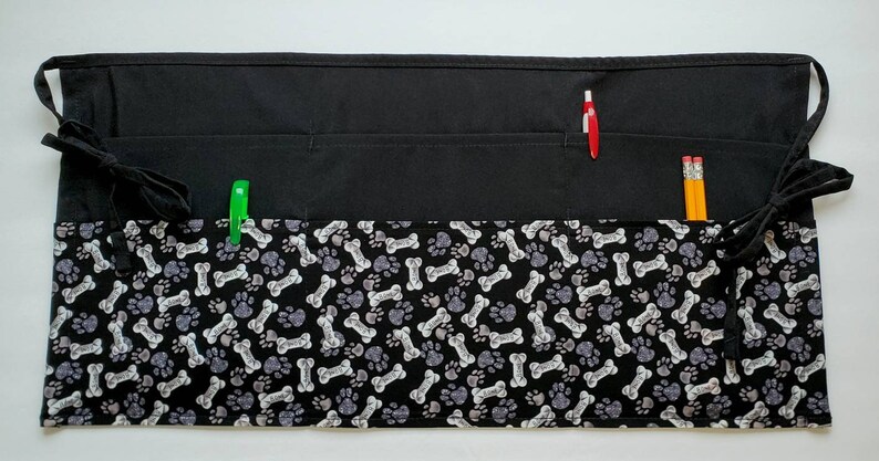 K&ouml;nnte beinhalten: Schwarze Sch&uuml;rze mit mehreren Taschen und einem Muster aus Hundeknochen und Pfoten. Die Sch&uuml;rze hat zwei verstellbare B&auml;nder.