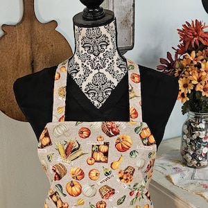 May include: A beige apron with a repeating pattern of pumpkins, squash, corn, and the word "pumpkins". The apron is displayed on a mannequin with a black shirt and a patterned neck. The background includes a wooden cutting board and a jar of buttons.