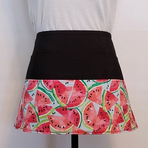 May include: A black apron with a watermelon print. The apron has two pockets on the front and a wide waistband.